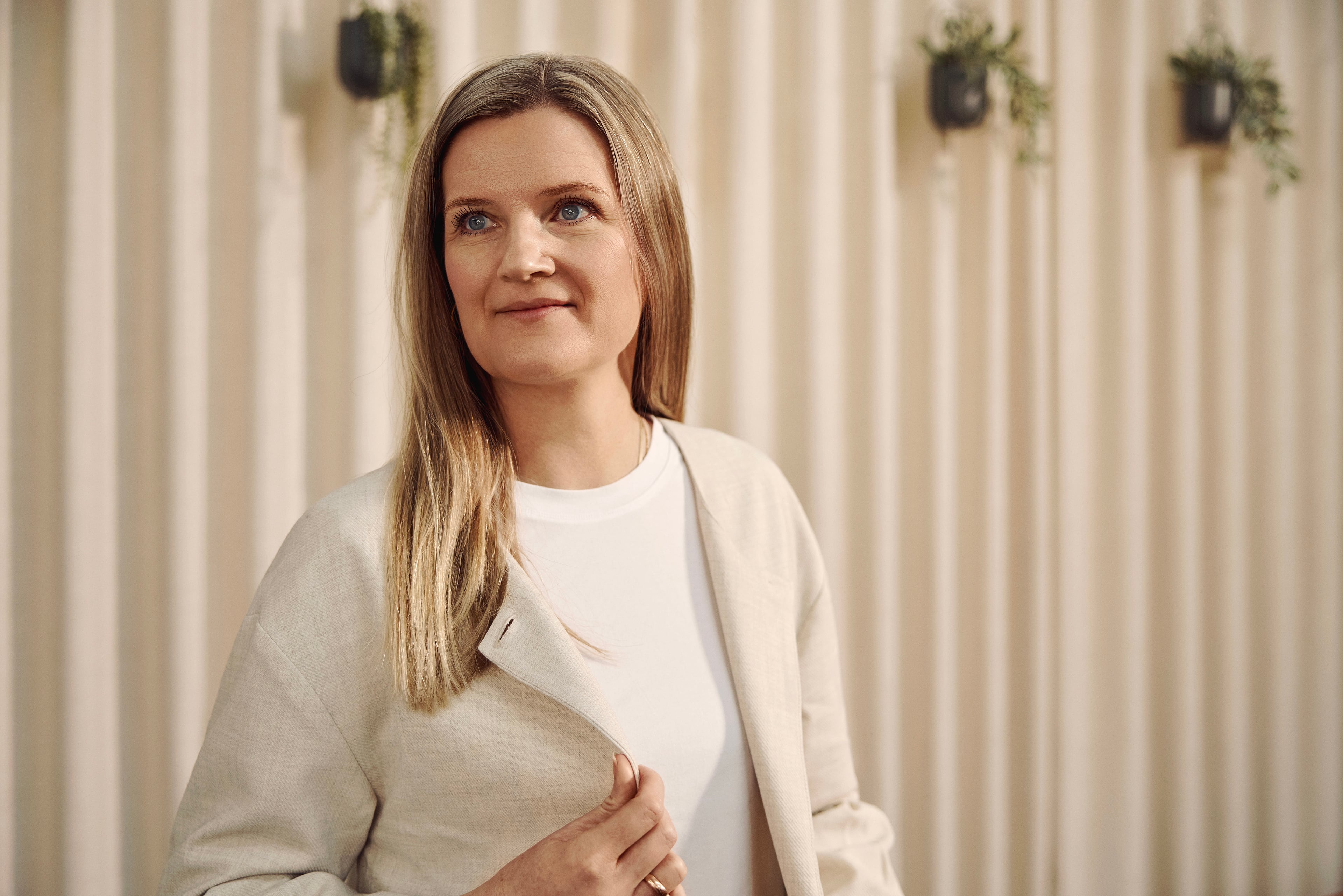 Kvinde i beige cardigan og hvid t-shirt foran stribet væg med hængende planter