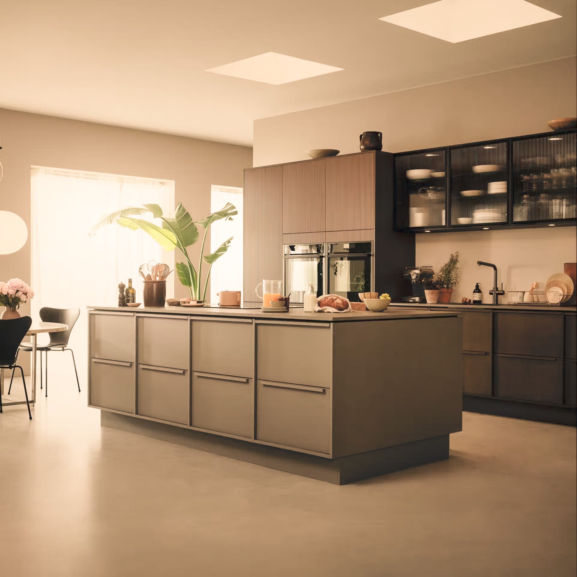 Moderne køkken med beige og sorte skabe, køkkøø med indbyggede ovne, grønne planter og naturligt lys fra vinduer