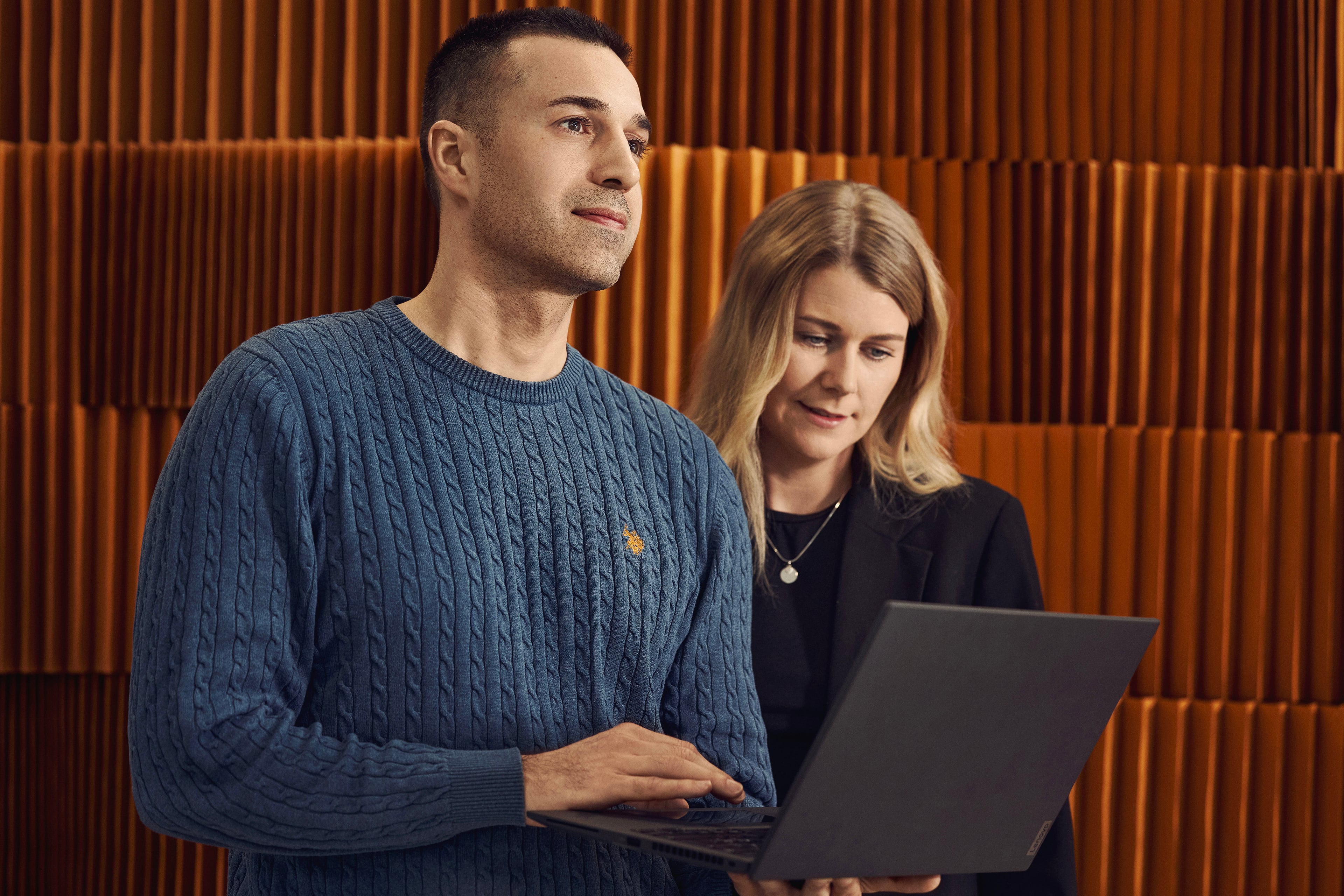 To personer arbejder sammen ved en bærbar computer foran orange vægpanel